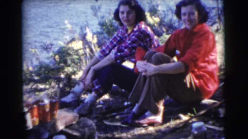 Two women sit together beside a small lakeside campfire with cans and supplies in 1956 Miami-area 8mm home-movie footage.