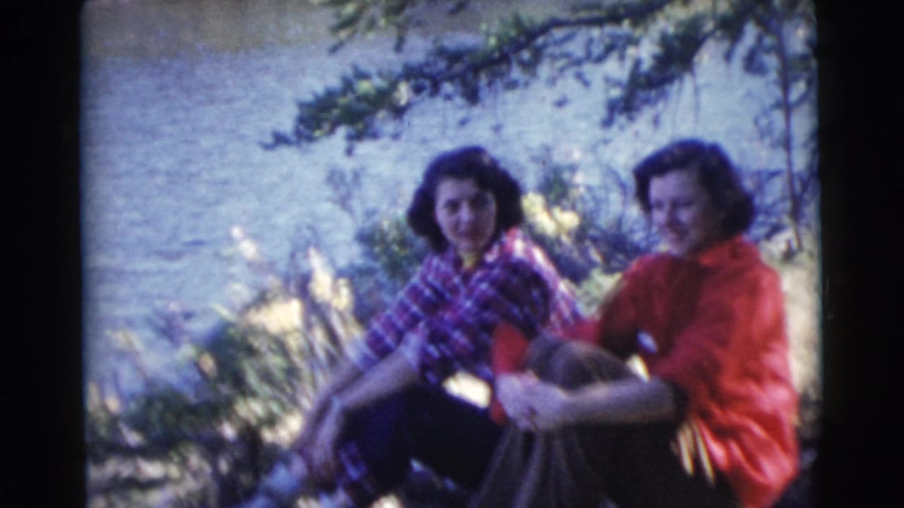 Two women sit near a lake under tree shade, one in plaid and one in a red top.