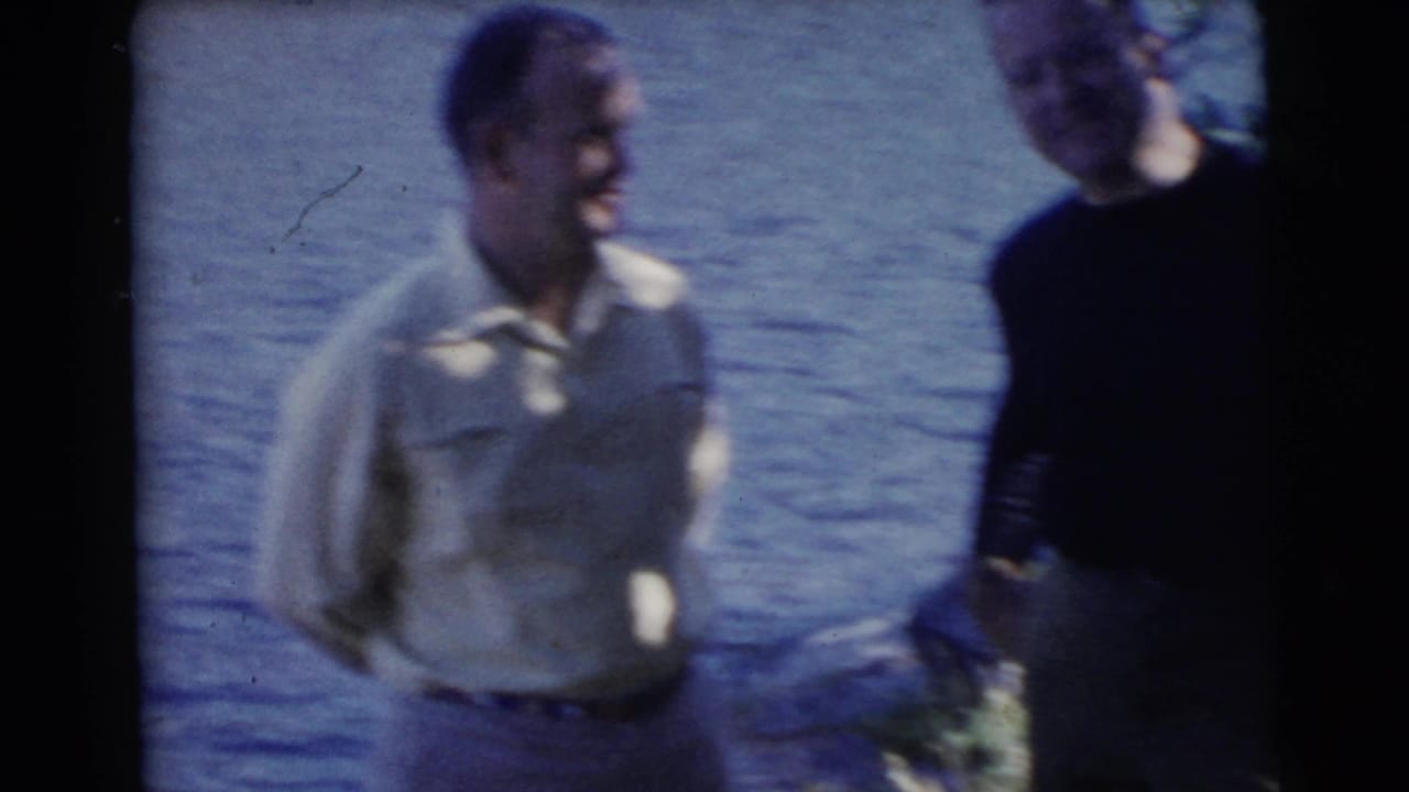 Two men stand near the water, one in a light shirt and one in a dark sweater.