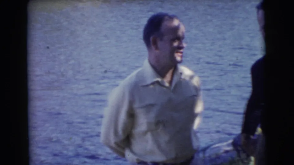 A man in a light collared shirt stands smiling beside the Miami lake, with another adult partly cropped at frame edge.