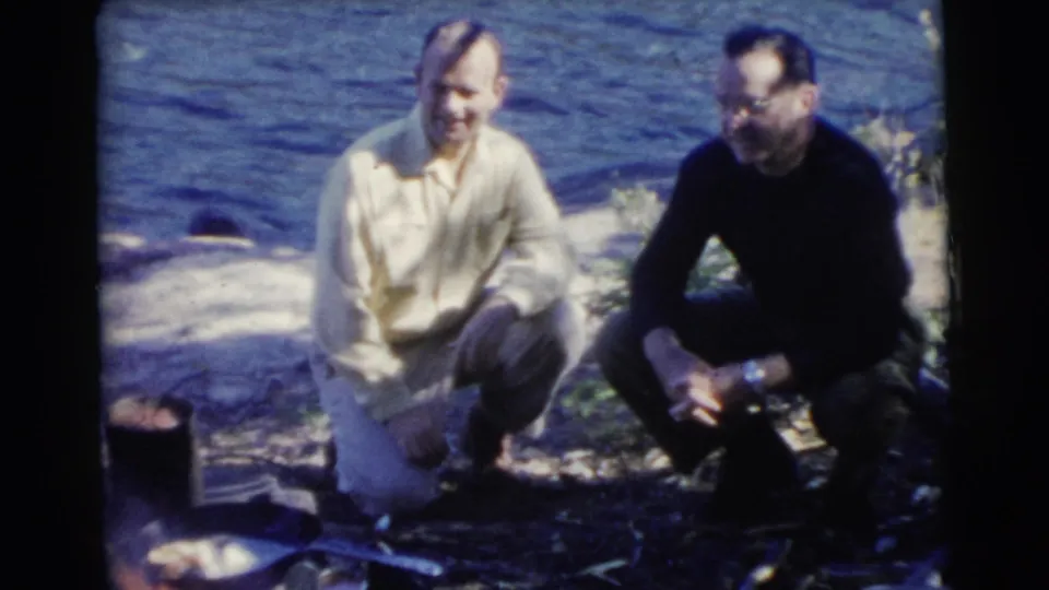 Two men in 1950s casual clothing crouch by a Miami lakeshore, with shaded ground and small outdoor items nearby.