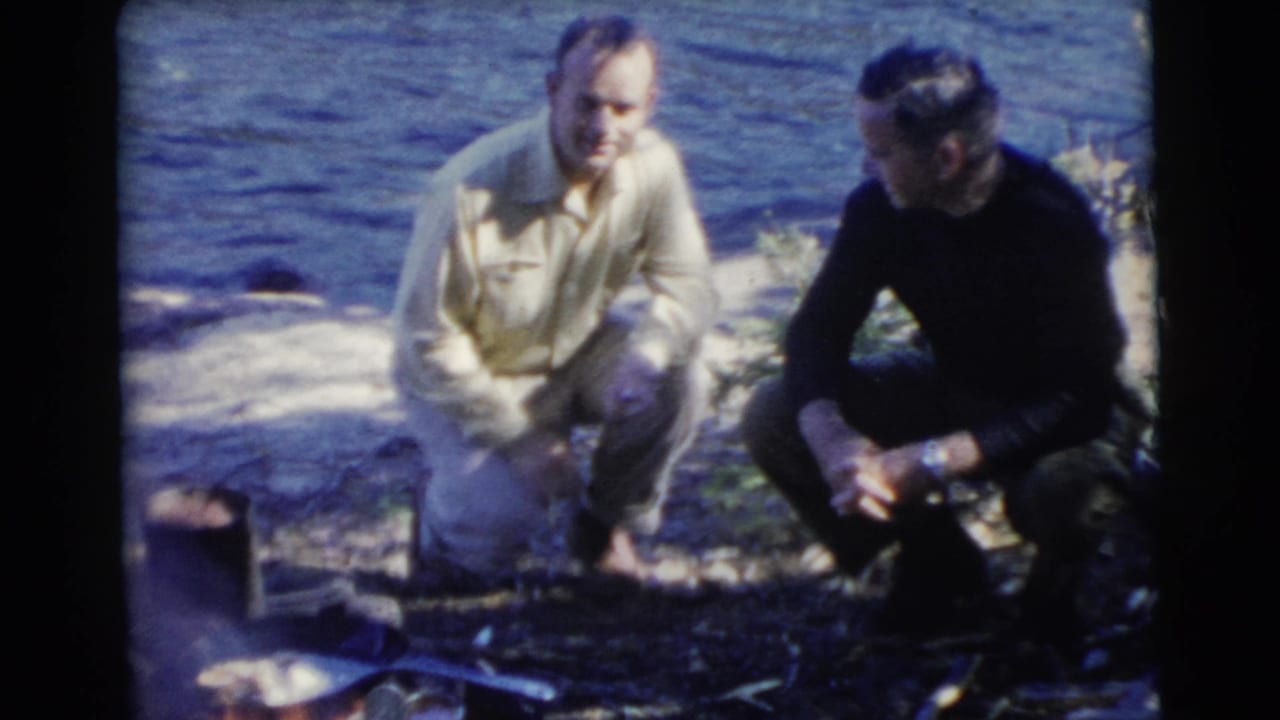 Two men crouch beside a small shoreline fire with lake water behind them.