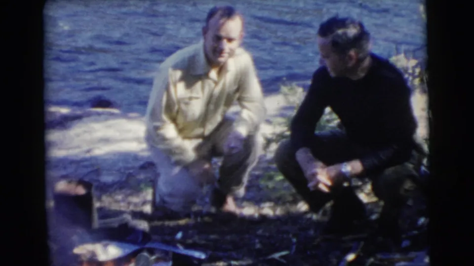 In 1956 Miami-area home-movie footage, two adult men crouch near a small campfire at a lakefront shoreline.