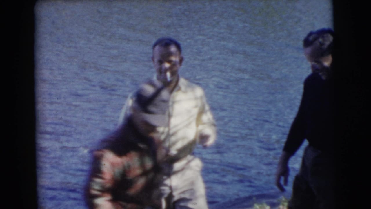 A man in light 1950s casual clothing stands at the lakefront as another adult bends near the shoreline.