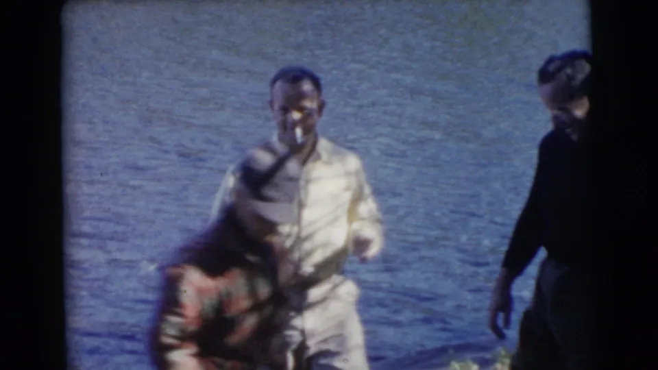 A man in light 1950s casual clothing stands at the lakefront as another adult bends near the shoreline.
