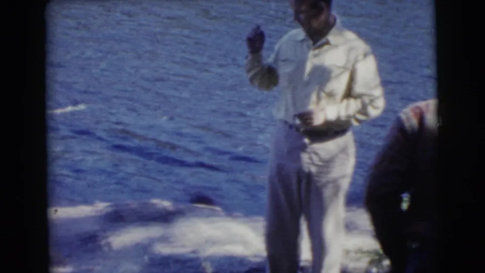 The light-clothed man stands on the lakefront shoreline, raising a hand as another adult is partly visible nearby.