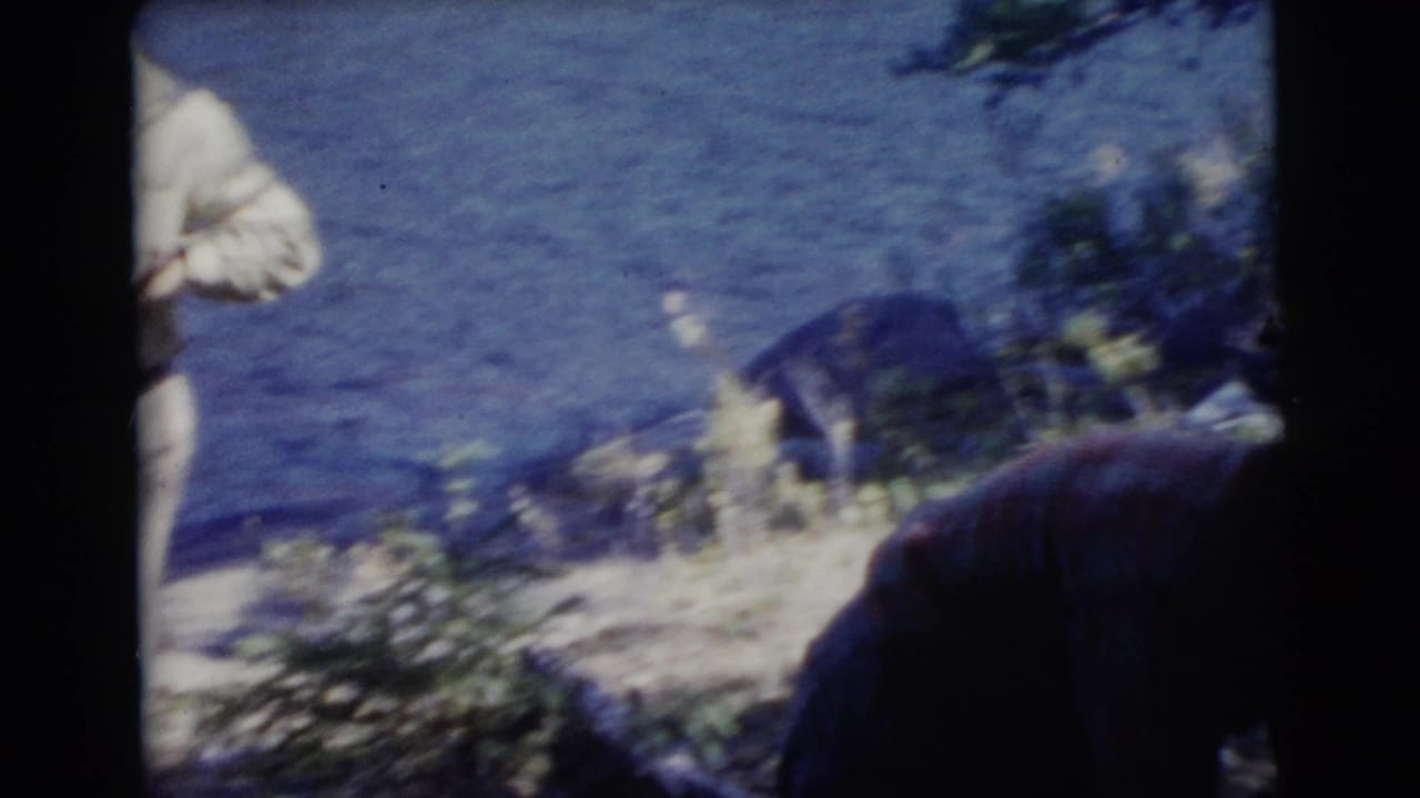 Adult figures near a shaded riverbank with water, rocks, and sparse plants visible.