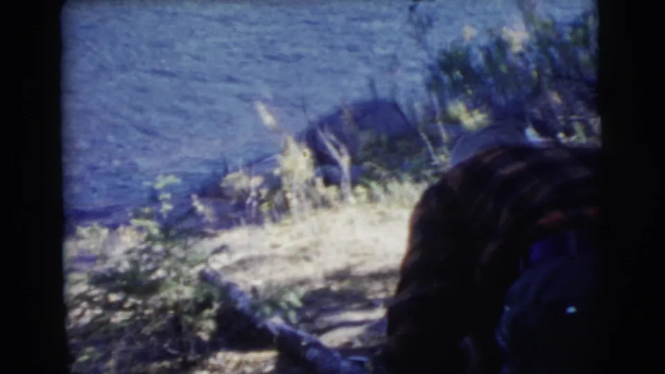 1956 Miami home-movie frame showing a bent-over adult in the foreground near a rocky riverbank and blue-toned water.