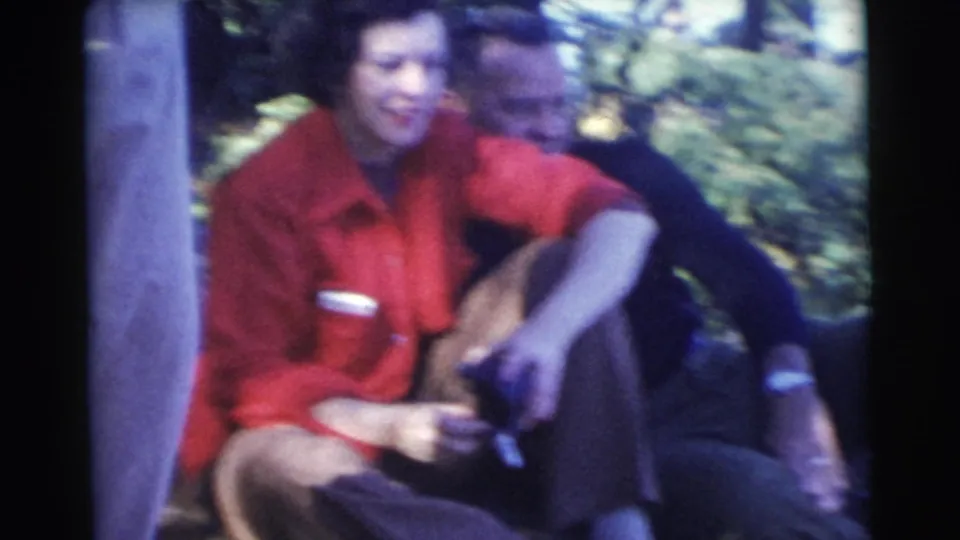 A woman in a red shirt sits beside a man in dark clothing outdoors, with trees and shaded daylight behind them.