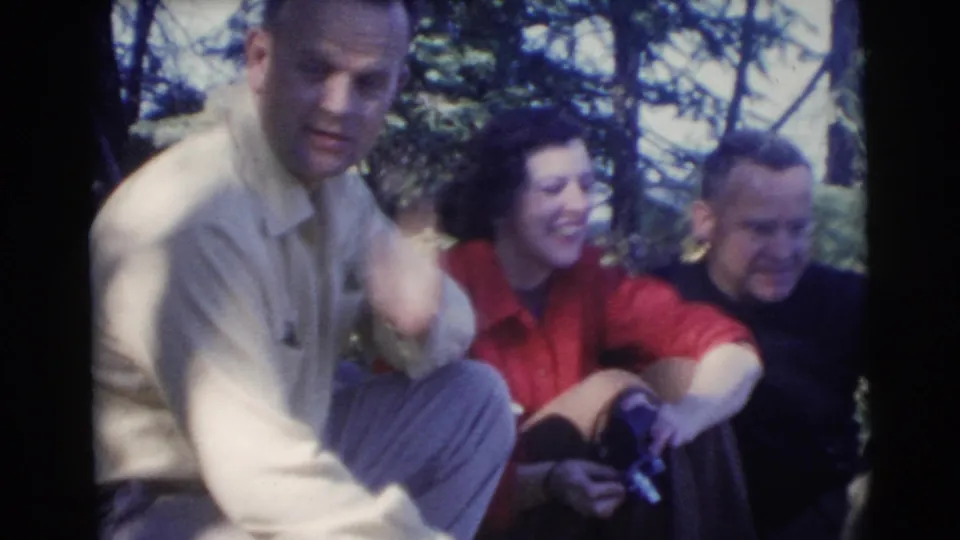Three adults sit together under trees in 1956 Miami home-movie footage, with one woman smiling and two men nearby.