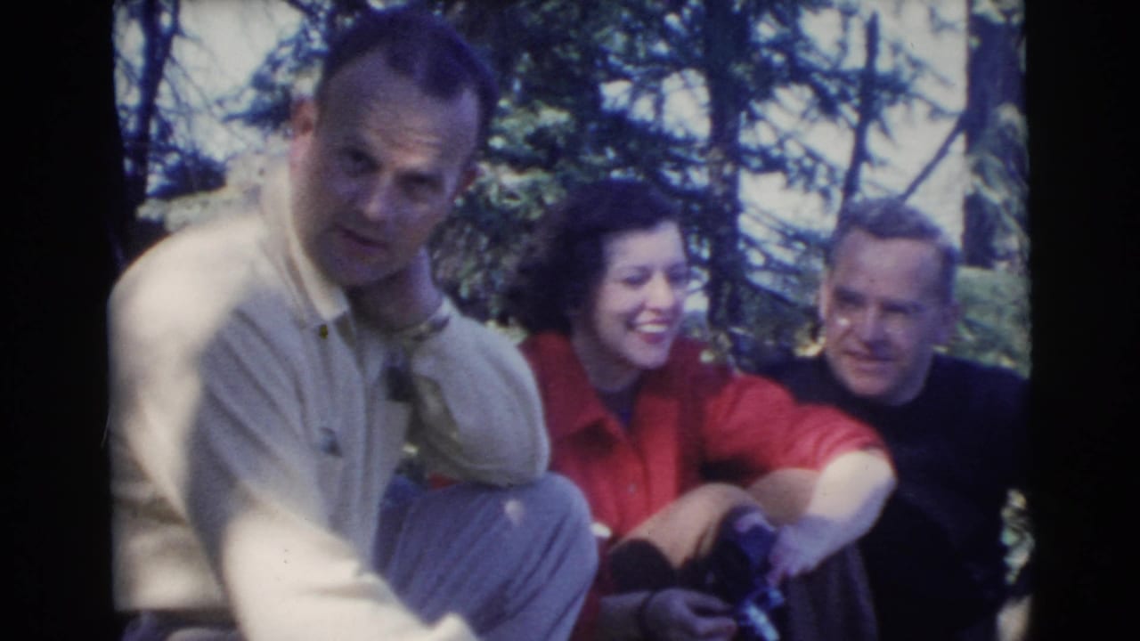 Three adults seated outdoors in wooded shade, with one man holding a cigarette.