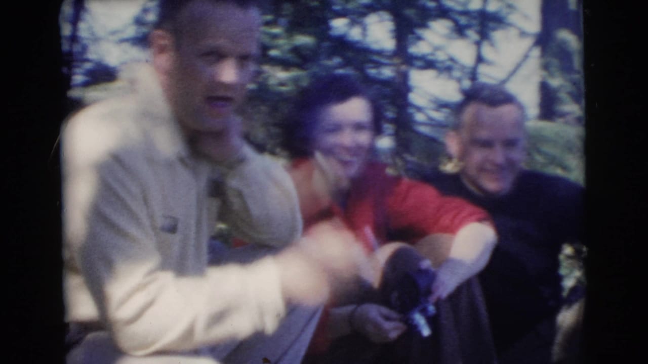 1956 Miami home-movie frame of adults relaxing in wooded shade while a man in a light shirt gestures with a cigarette.