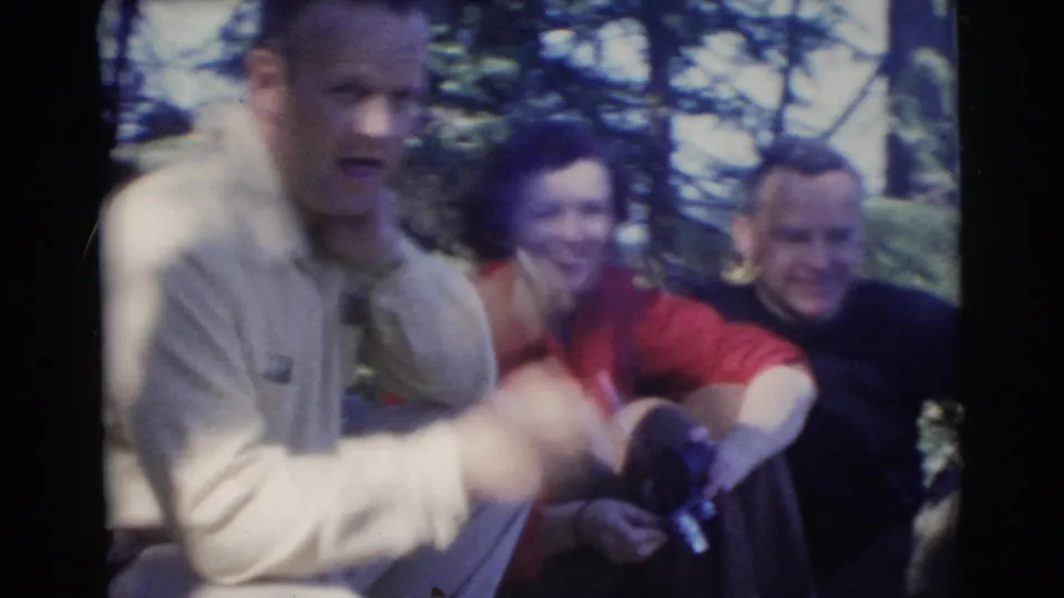 1956 Miami home-movie frame of adults relaxing in wooded shade while a man in a light shirt gestures with a cigarette.