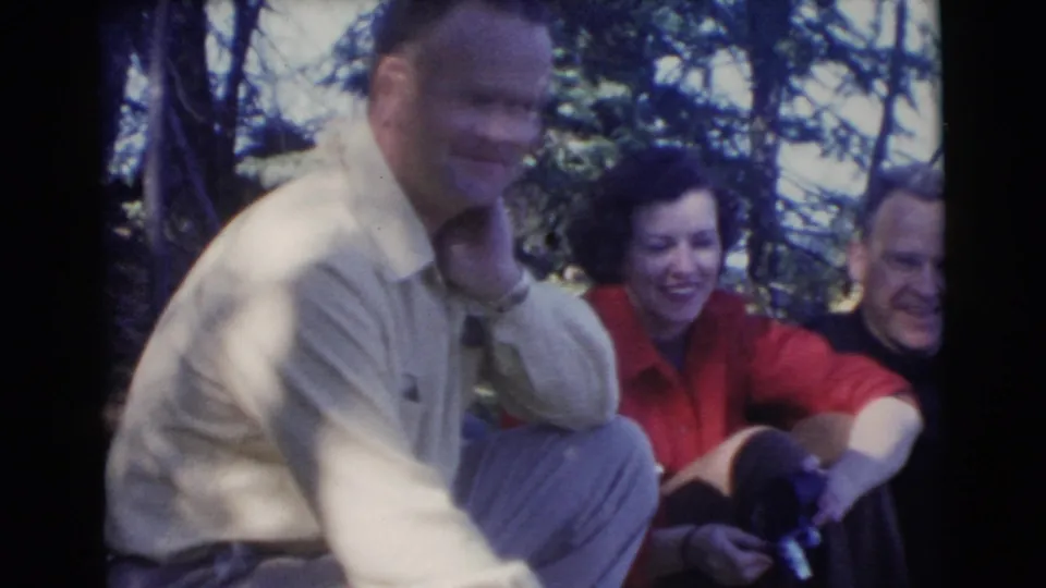 1956 Miami home-movie frame showing three adults smiling together in a shaded wooded outdoor setting.