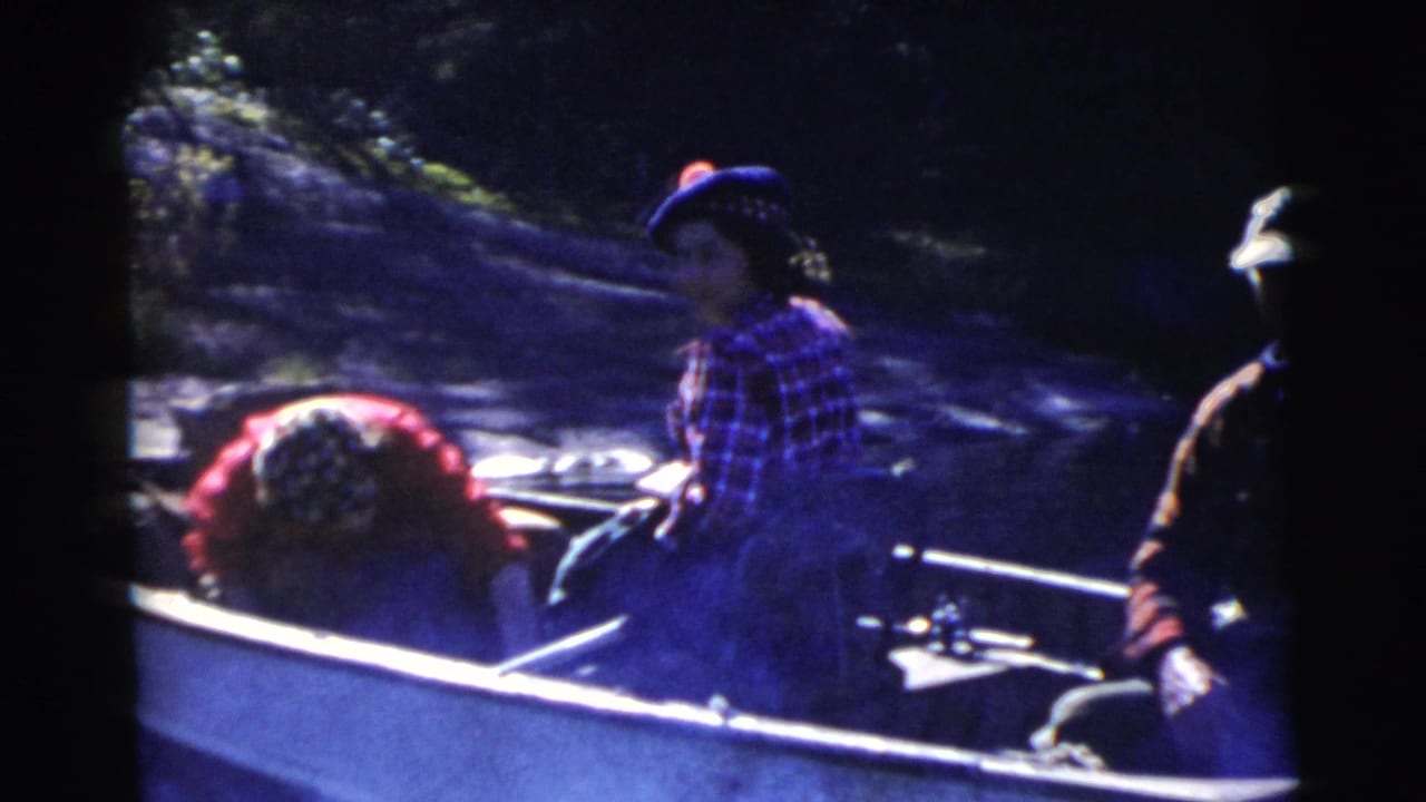 Adults seated in a small rowboat on a lake, with oars and a wooded bank behind them.