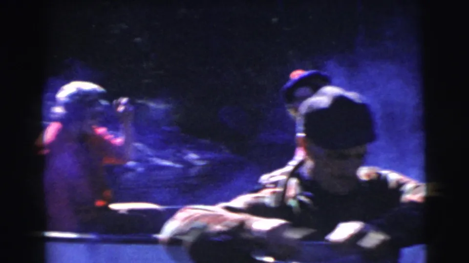 Home-movie frame of rowboat passengers in shadow, with a cap-wearing man in the foreground and hazy lake light behind.