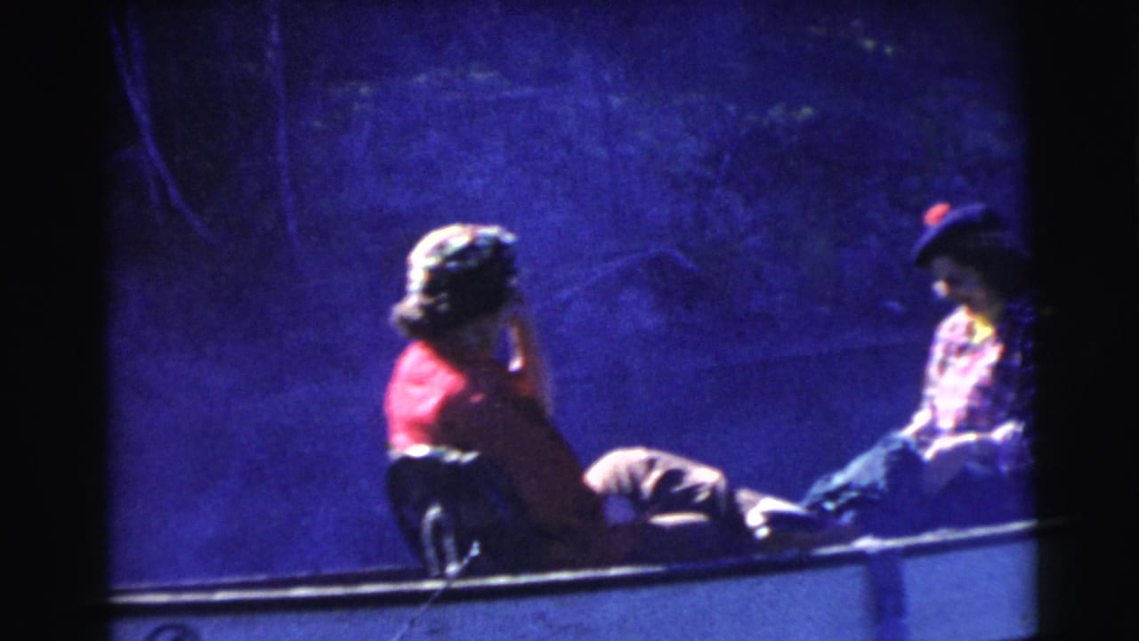 1956 Miami home-movie frame showing two seated passengers in a small boat, with a woman in red near the bow and another passenger opposite.
