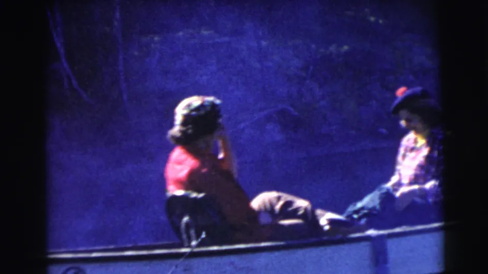 1956 Miami home-movie frame showing two seated passengers in a small boat, with a woman in red near the bow and another passenger opposite.