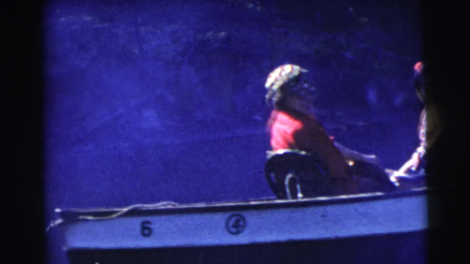 1956 Miami home-movie frame with a red-clad passenger seated in a small motorboat, its side number partly visible in blue shadow.