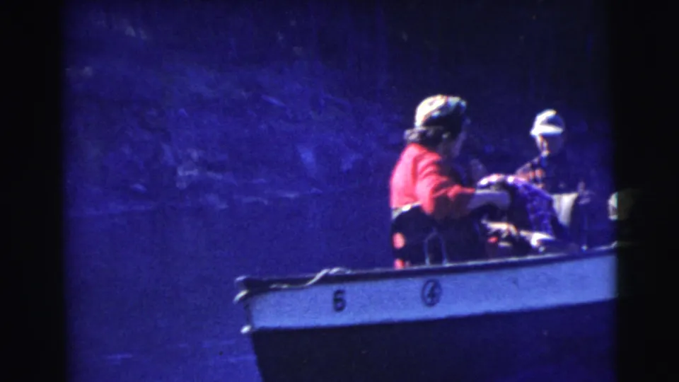 1956 Miami home-movie frame showing multiple seated passengers in a small numbered boat, including hats and patterned clothing in deep blue light.