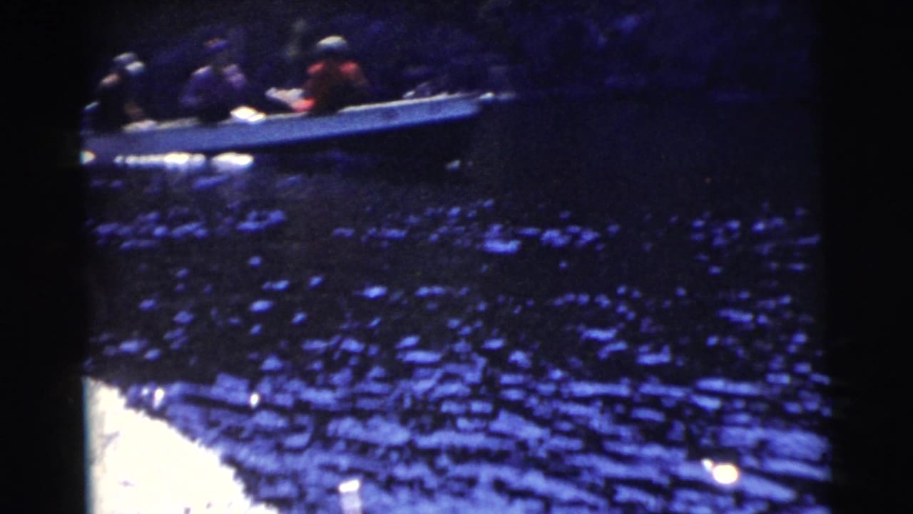 1956 Miami 8mm frame showing a small open motorboat with passengers on dark sunlit water near a wooded bank.