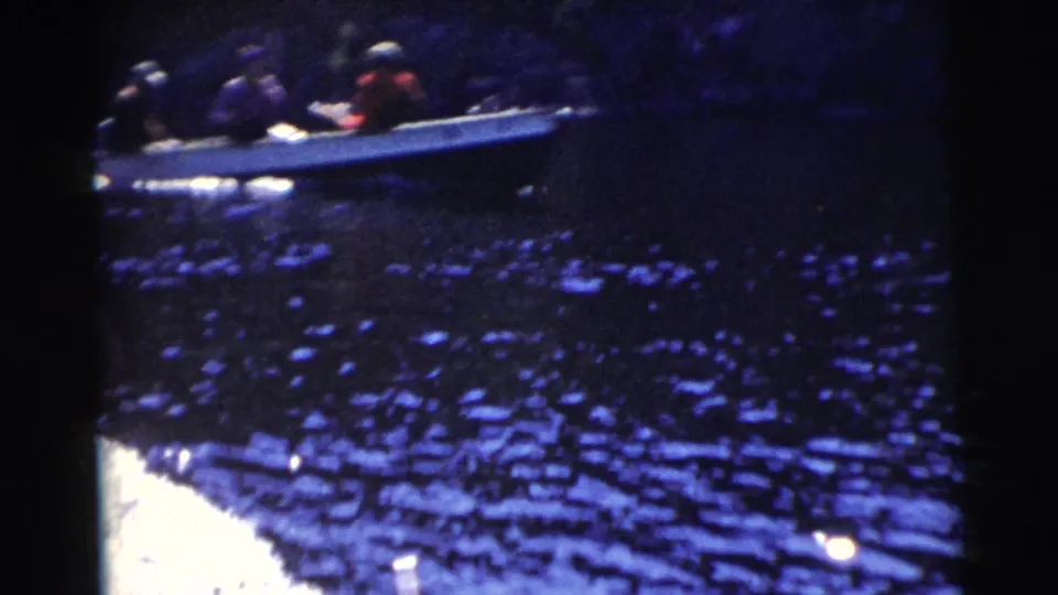 1956 Miami 8mm frame showing a small open motorboat with passengers on dark sunlit water near a wooded bank.