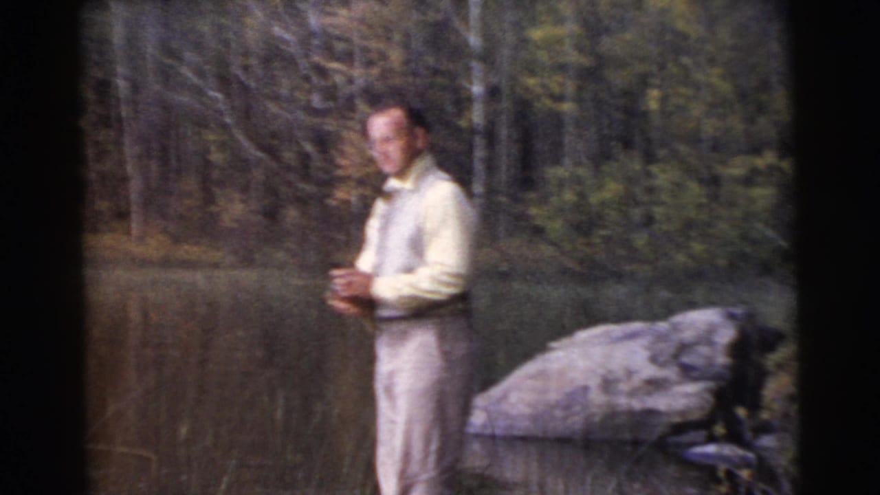 Man in light clothing stands beside a wooded lake holding fishing gear.