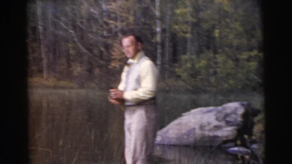In 1956 Miami home-movie footage, a man in light clothing stands near a calm wooded lake with a large shoreline rock nearby.