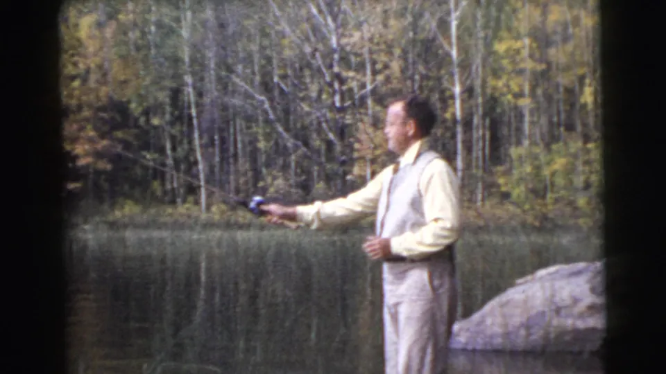 A 1950s 8mm frame shows the man extending his fishing rod over the lake, with wooded shoreline and muted foliage behind him.