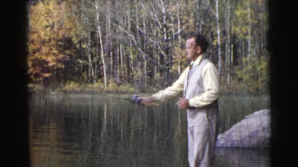 The man holds the rod outward beside the calm lake, framed by trees, shoreline rocks, and warm mid-century 8mm color.