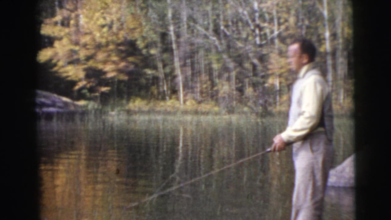 Man standing by calm water with a long fishing rod and trees behind him.