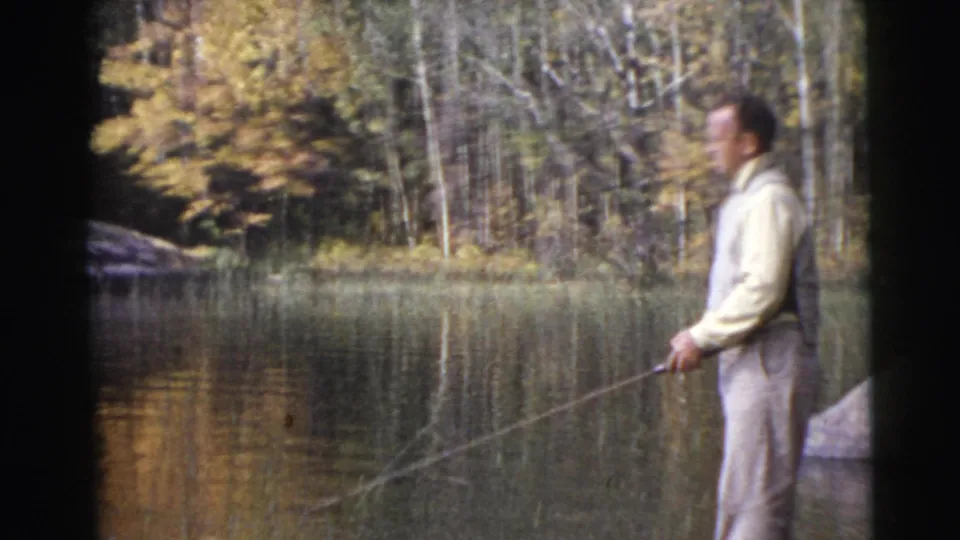 1956 Miami home-movie frame of an adult man in light clothing fishing beside calm water with trees across the bank.
