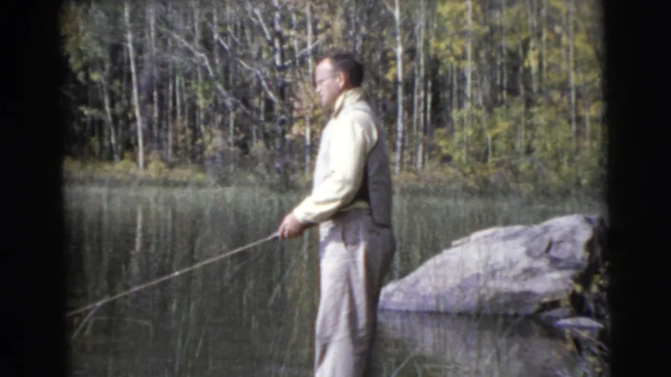 Side view of the man fishing beside a calm pond, with rocks, reeds, and a wooded bank visible in the 1956 Miami footage.