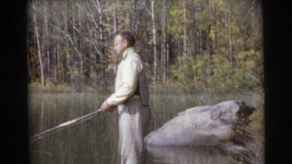 The man holds his fishing rod near the water, framed by shoreline rocks and trees in the vintage Miami home-movie scene.