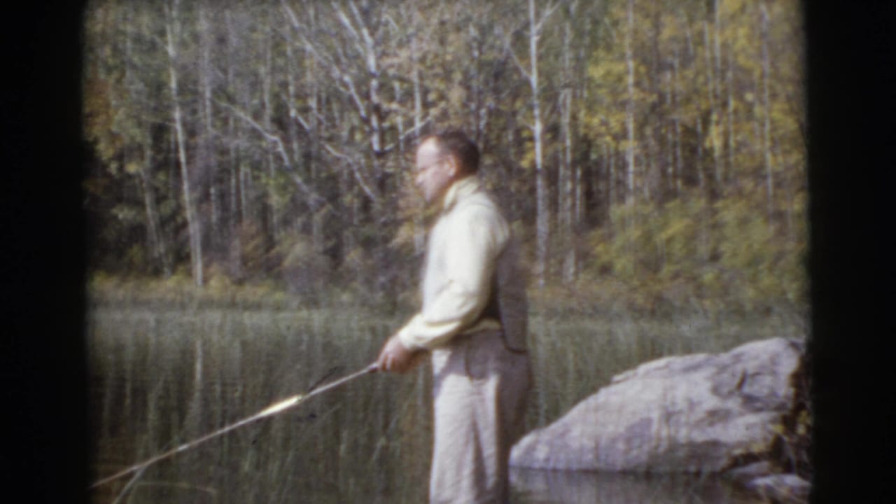 Man in light clothing stands near a lake holding a fishing rod beside a large rock.