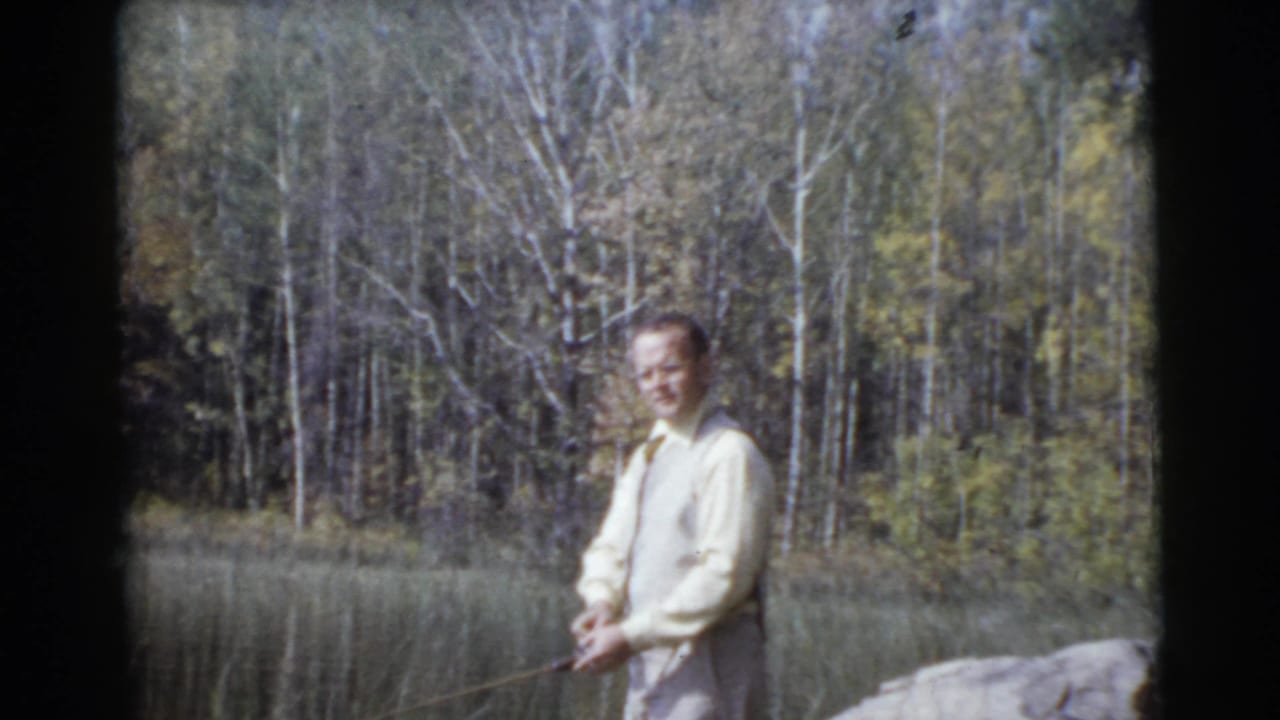 Man faces the camera while holding a fishing rod in front of a wooded lakeshore.
