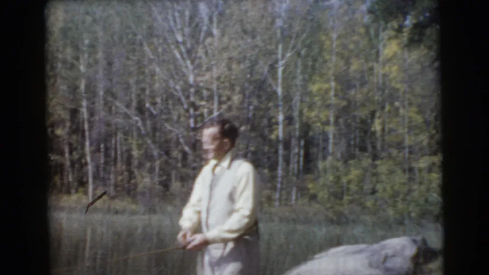 A light-clothed man stands by the lake with a fishing rod, framed against trees and water in metadata-supported 1956 Miami home-movie footage.