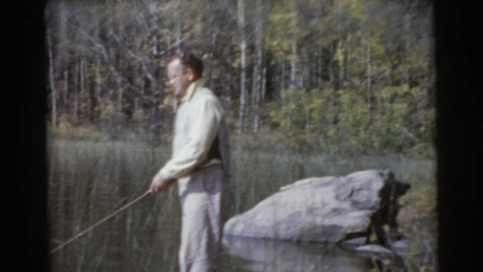 Side view of the fisherman reeling near a large lakeside rock, with wooded shoreline behind him in 1956 home-movie footage identified as Miami.