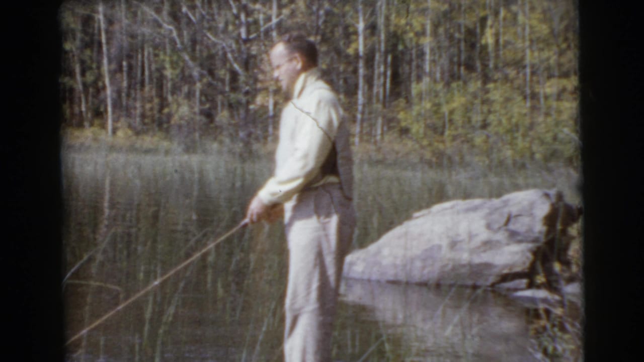 In metadata-supported 1956 Miami-area home-movie footage, a man in light clothing stands beside a calm lake holding a long fishing rod.