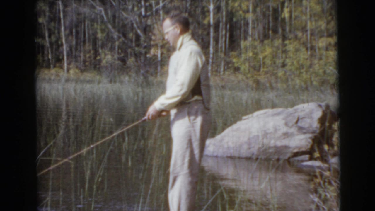 The man is shown in profile at the edge of a calm lake, holding his fishing rod toward reeds in 1956 home-movie footage.