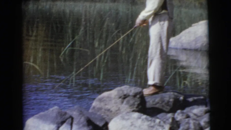 A closer lower-frame view shows the man standing on shoreline rocks with his long rod angled over the water.