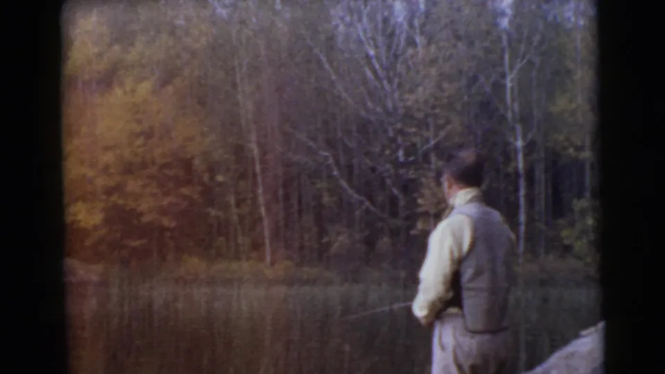 A rear view shows the man fishing beside the lake, with reeds, rocks, and autumn-colored woodland visible across the water.