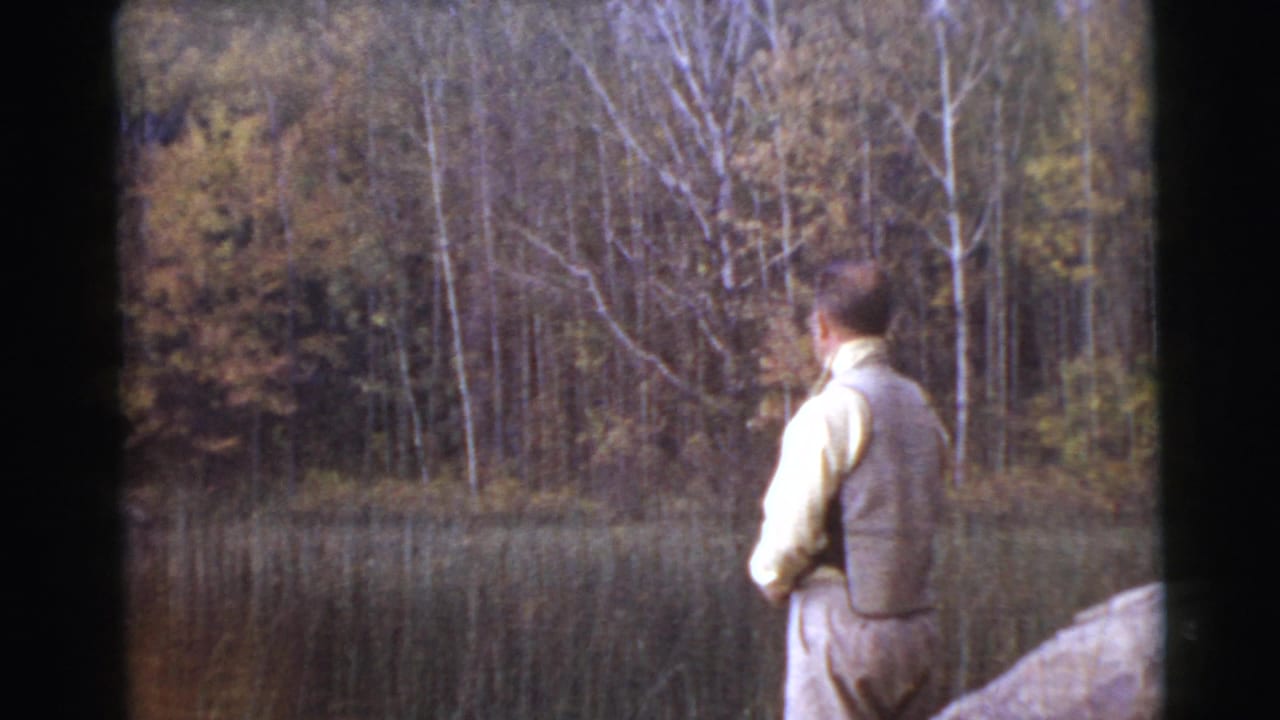 Man in a vest stands near a wooded lake holding a fishing rod.
