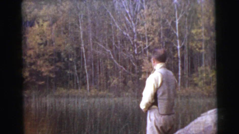 1956 8mm frame of an adult man fishing beside a calm wooded lake, with reeds and autumn-toned trees in the background.