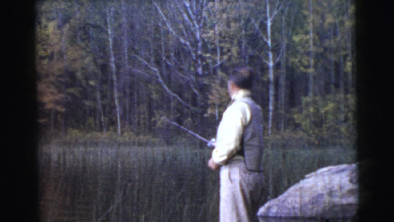 Man fishes from a rocky lakeside edge with trees across the water.