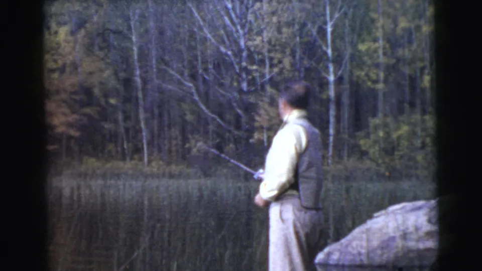 Adult man in light clothing fishes at the edge of a calm lake in metadata-listed Miami, with reeds and wooded shoreline beyond.