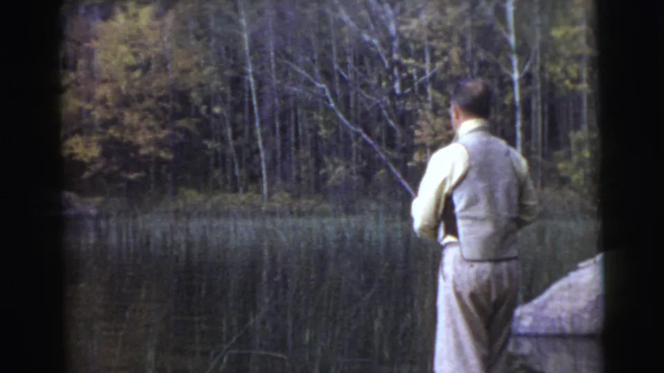 Rear view of the fisherman holding his rod near still lake water, framed by reeds, rocks, and a wooded shoreline.