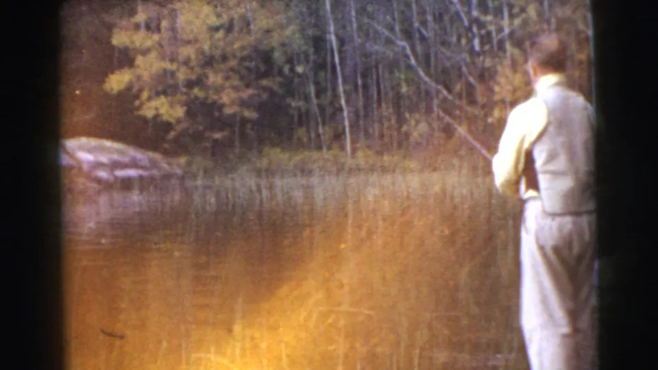 The fisherman appears at the right edge of the frame beside golden lake reflections, reeds, rocks, and autumn-colored trees.