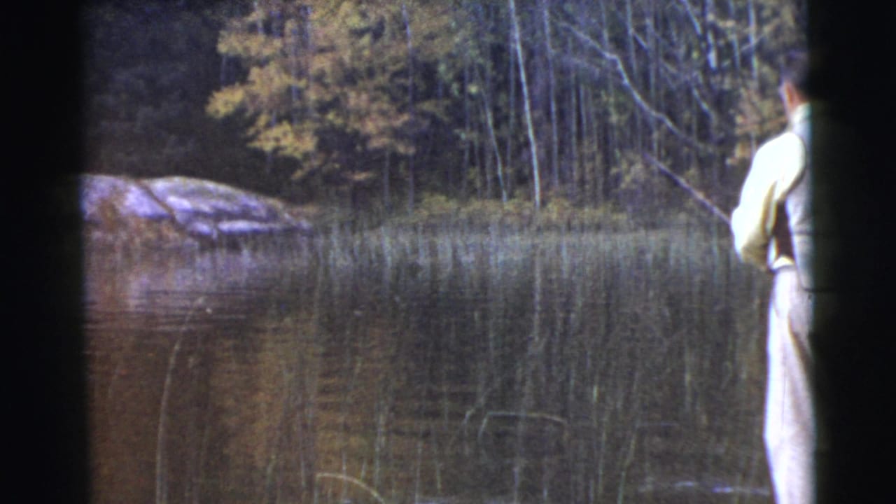 1956 8mm frame from Miami metadata showing a man from behind fishing at a wooded lake edge with reeds, rock, and autumn-toned trees.