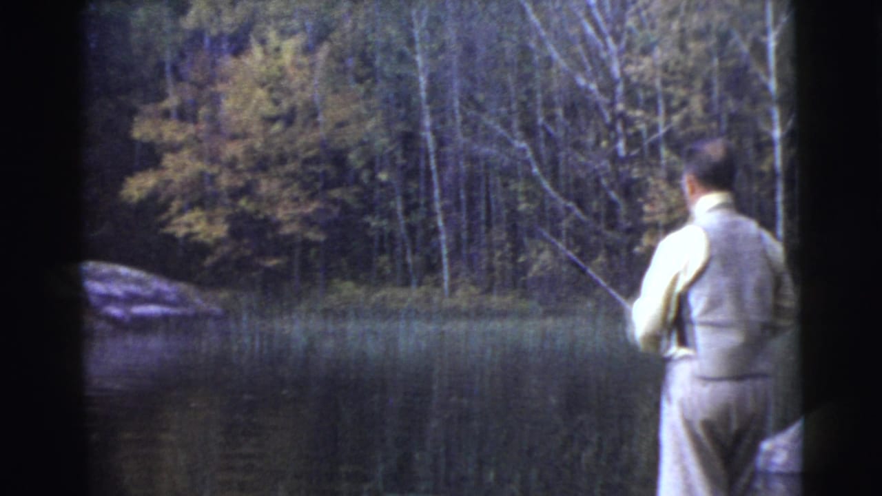 1956 8mm frame from Miami metadata showing a man in a vest fishing from a lakeshore, with calm water, reeds, trees, and a shoreline rock.